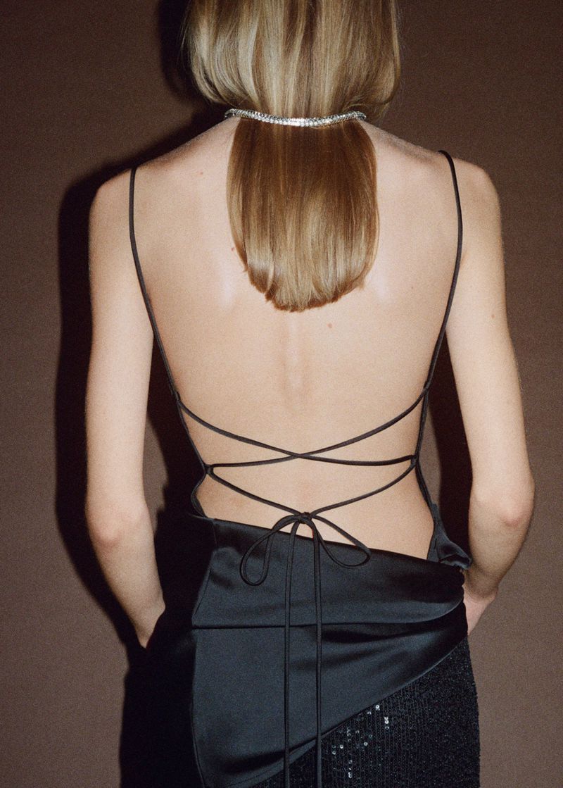 Back view of a woman with fair hair wearing a strappy black dress with a sequin skirt against a dark brown background in a studio.