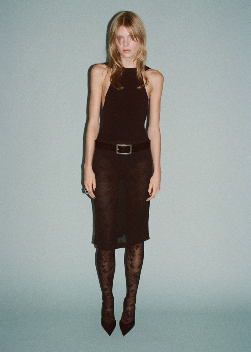 Full-length shot of a slender woman in a black halter top, patterned midi skirt, floral tights, and pointed shoes against a light blue backdrop.