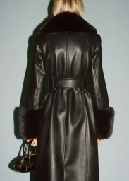 Model seen from behind wearing a black faux fur-trimmed coat and holding a handbag against a plain teal wall.