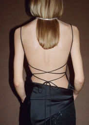 Model seen from behind wearing a backless black dress with spaghetti straps and a rhinestone choker against a plain brown backdrop