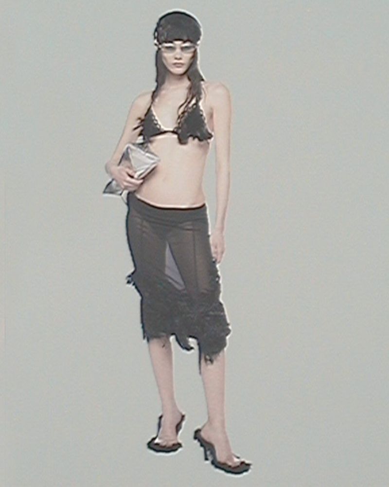 A woman poses in a black bikini top and sheer skirt, holding a silver bag, wearing translucent glasses and black heels against a grey background.