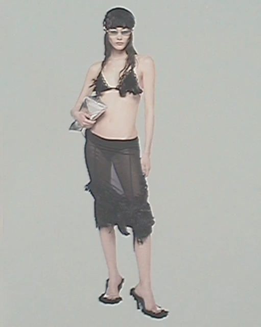 A woman poses in a black bikini top and sheer skirt, holding a silver bag, wearing translucent glasses and black heels against a grey background.