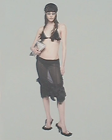 A woman poses in a black bikini top and sheer skirt, holding a silver bag, wearing translucent glasses and black heels against a grey background.