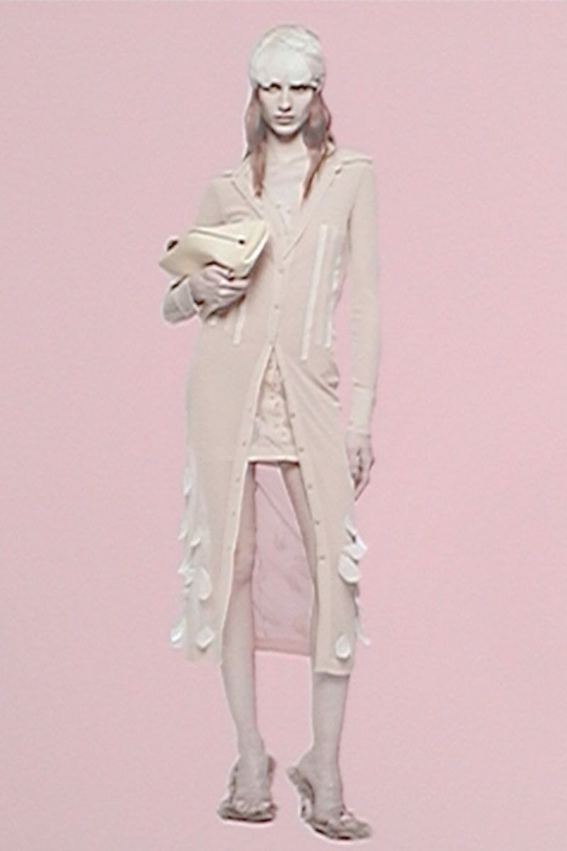 A woman in a pale pink sheer coat and sculptural hat holds a cream-colored clutch, standing on a pale pink background in a fashion editorial.