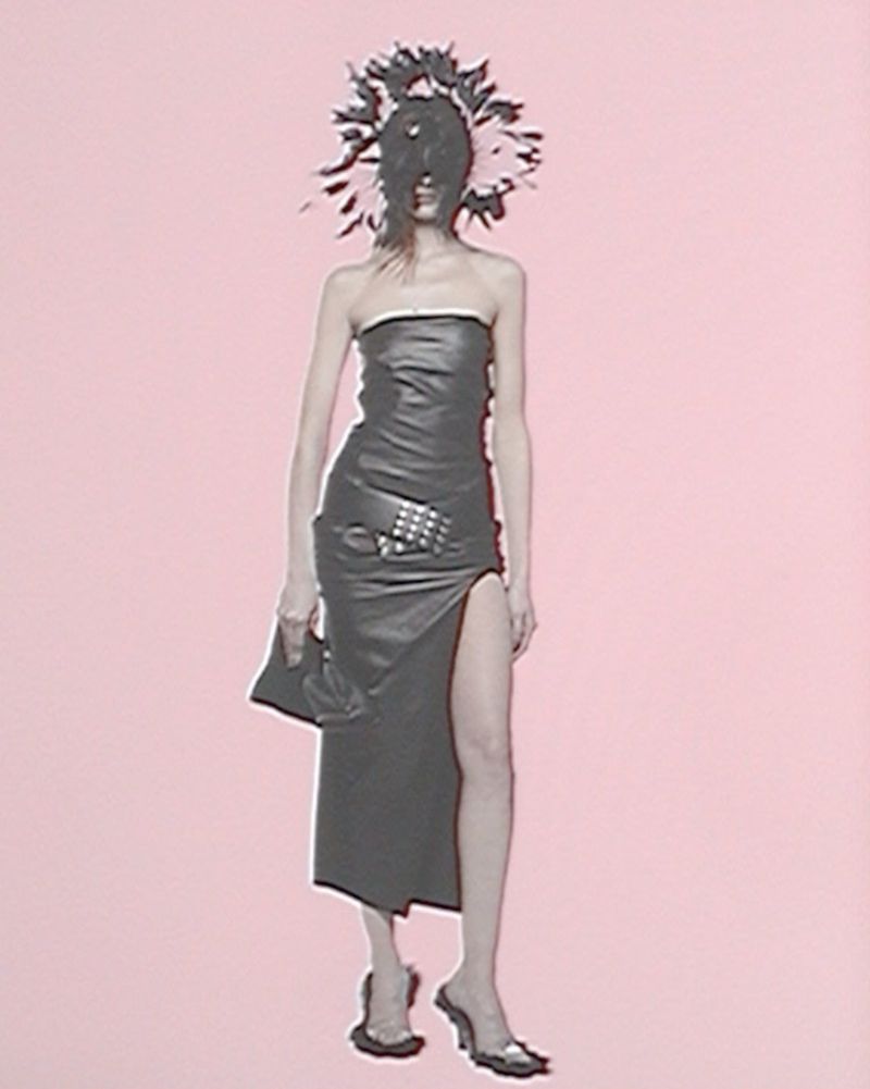 A fashion model wearing a black dress with a decorative headpiece stands in front of a pink backdrop, presenting an editorial look.