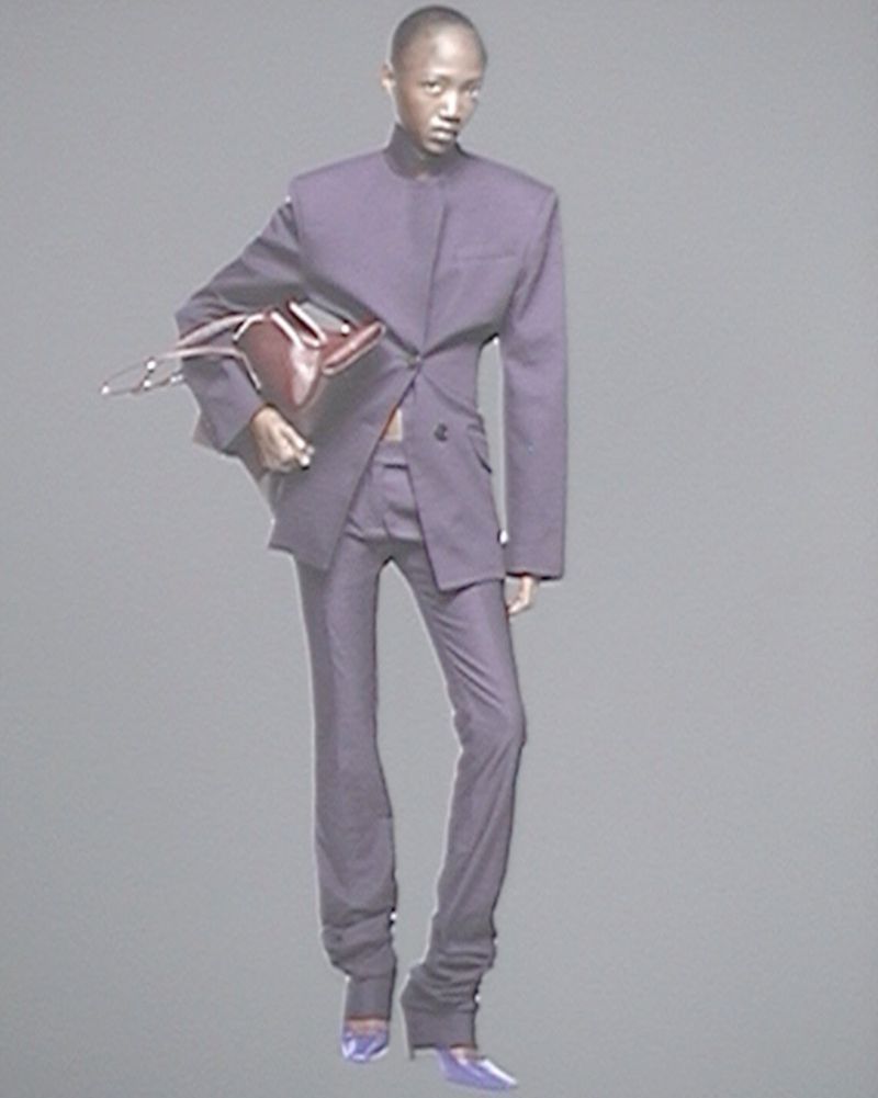 Fashion photo of a Black model in a purple suit holding a brown purse, set against a neutral background in a studio environment.