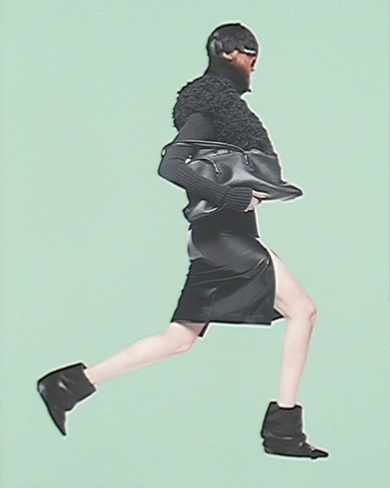 An adult fashion model is leaping forward while holding a handbag in front of a solid pale-green backdrop inside a photo studio.