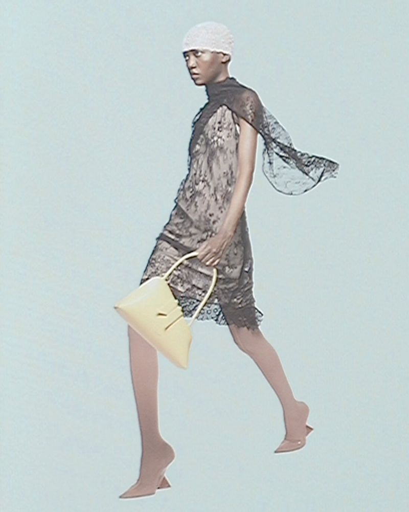An adult woman in a light-colored dress, and nude heels, walking with a purse against a light blue background in a studio fashion shot.