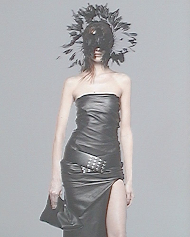 A slender fashion model presents a runway look with a strapless leather dress, matching clutch, feather headdress against a gray backdrop.