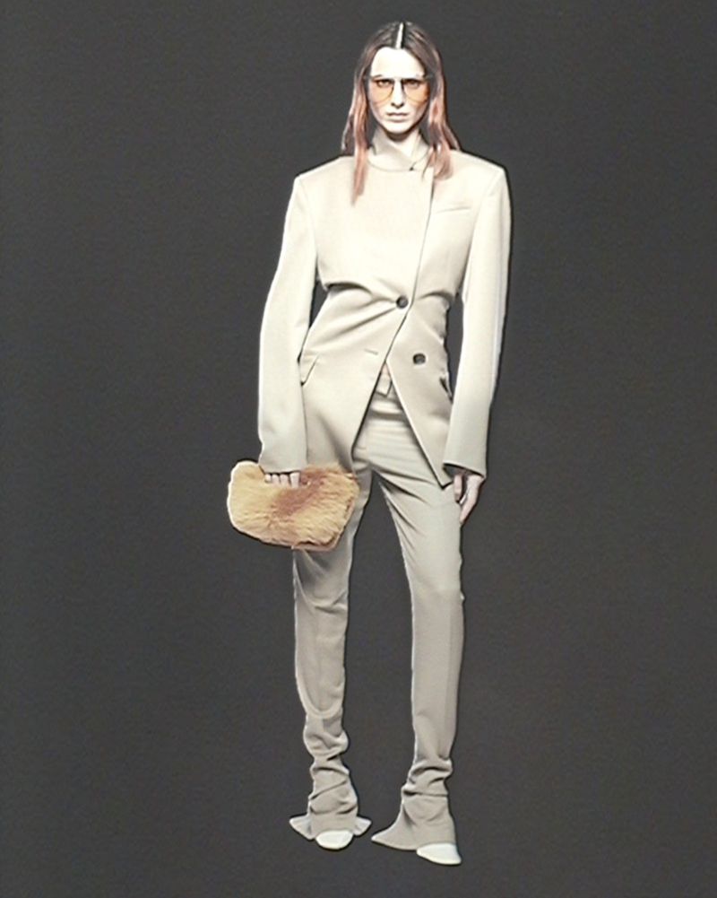 Fashion shot of a woman in a beige blazer and pantsuit holding a fur clutch against a dark grey background in a studio setting.