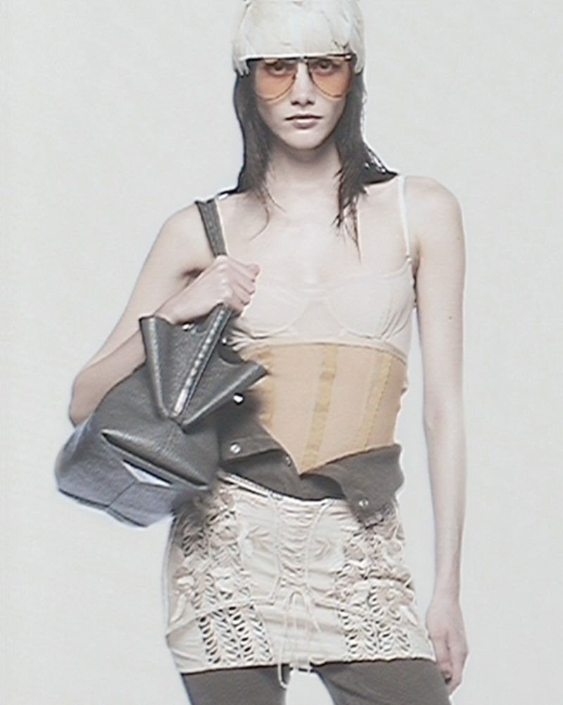 A runway model poses in a light-colored, translucent top and a beige corset, complemented by aviator sunglasses and a sculptural bag.