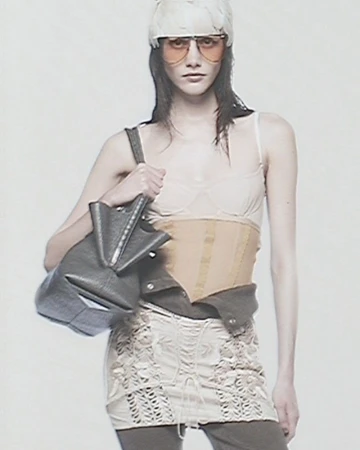 A runway model poses in a light-colored, translucent top and a beige corset, complemented by aviator sunglasses and a sculptural bag.
