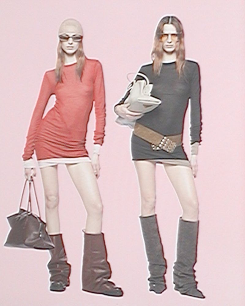 Two fashion models pose in front of a pink backdrop. One model has a handbag and sunglasses, the other a studded belt and purse.
