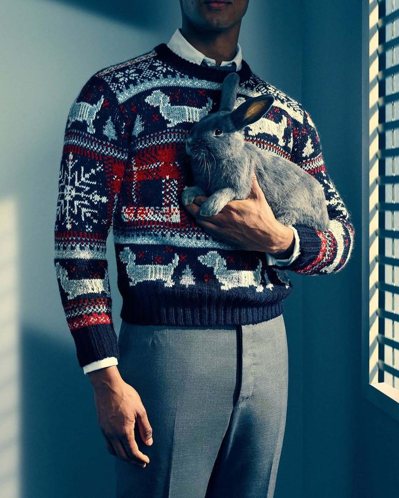 Man holding a gray rabbit, wearing a patterned winter sweater with dogs and snowflakes indoors, near window with blinds.