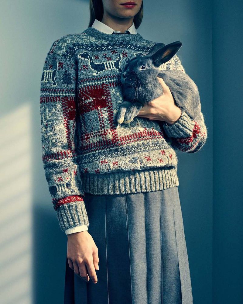 A woman wearing a patterned fair isle sweater and pleated skirt holding a dark rabbit against a grey wall