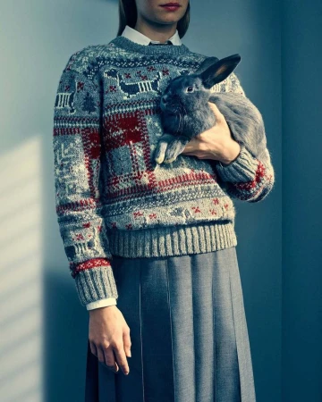 A woman wearing a patterned fair isle sweater and pleated skirt holding a dark rabbit against a grey wall