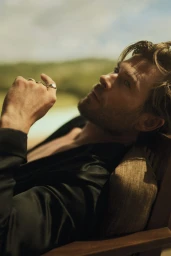 Chris Hemsworth seated in a chair outdoors wearing a dark shirt and silver rings with a soft sky and foliage background