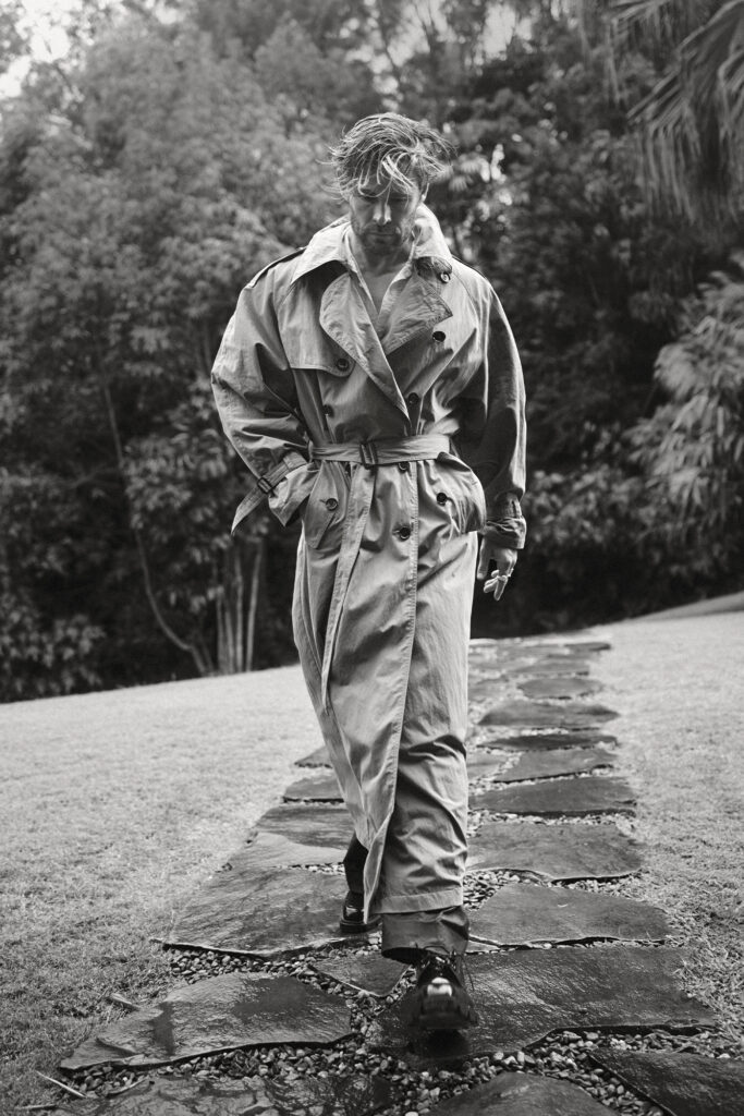 Monochrome portrait of a man in a trench coat walking on a stone path through a grassy, tree-filled outdoor setting in a stylish editorial.
