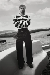 Chris Hemsworth standing with arms crossed wearing a striped polo shirt and dark trousers outdoors against an open landscape background