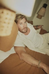 Chris Hemsworth reclining on a bed wearing a white t-shirt and open shirt, shot from an elevated foreground angle with warm lighting.