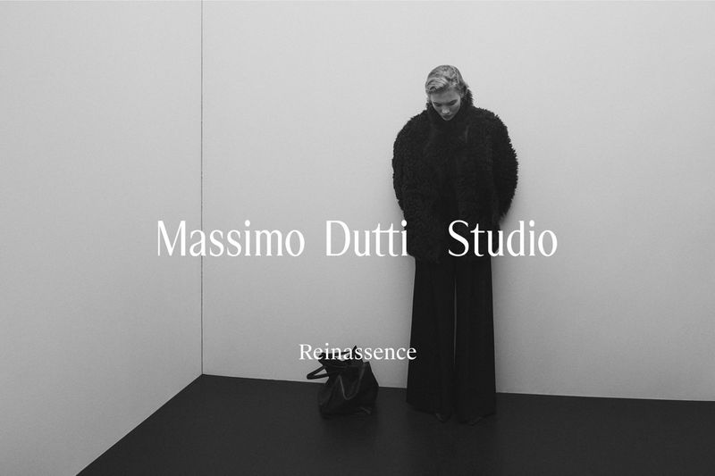 Fashion shot: A model poses indoors in a dark, textured coat and wide-leg pants in a minimalist studio setting, with a handbag.