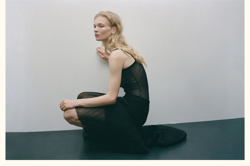 Woman with wavy blond hair is sitting in a black sheer dress, facing away from the camera in a simple indoor setting.