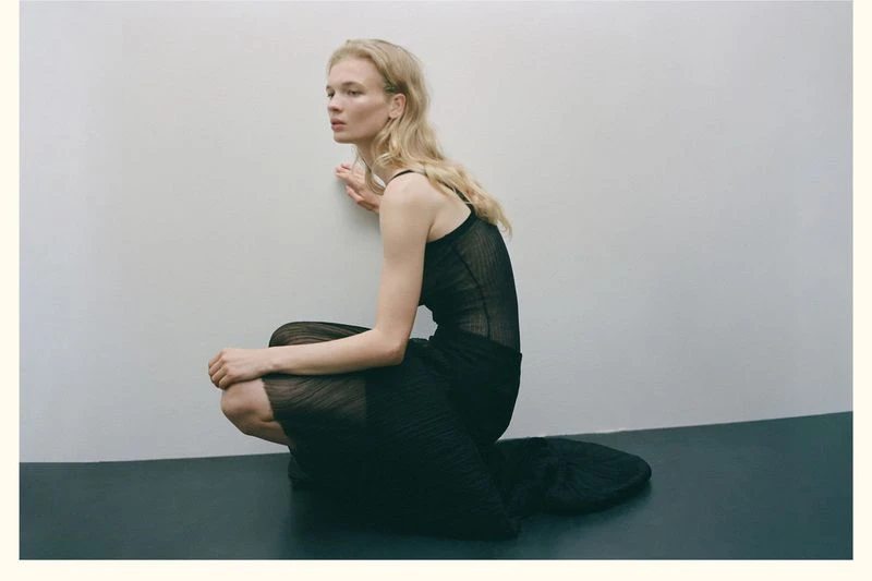 Woman with wavy blond hair is sitting in a black sheer dress, facing away from the camera in a simple indoor setting.