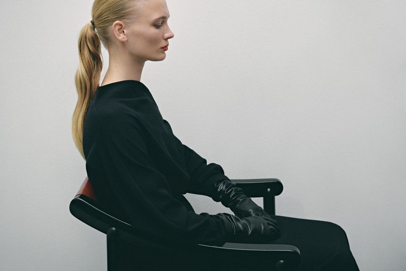 Side profile of a fair-skinned woman with a blonde ponytail, red lipstick, wearing a black dress and gloves, sitting in a black chair.