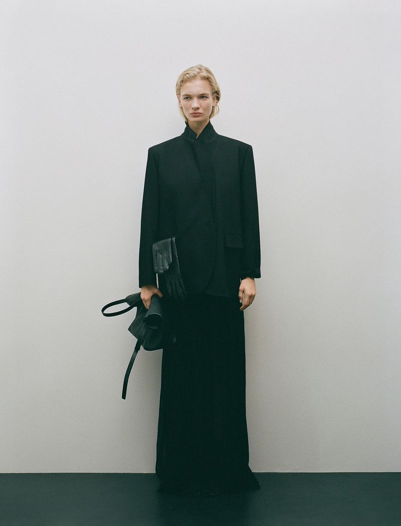 A woman in a black jacket and skirt stands in a minimalist setting, holding a black bag, creating a timeless, elegant fashion image.