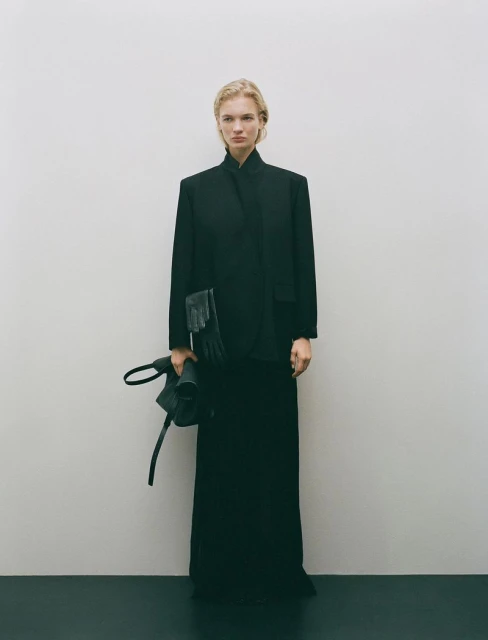 A woman in a black jacket and skirt stands in a minimalist setting, holding a black bag, creating a timeless, elegant fashion image.