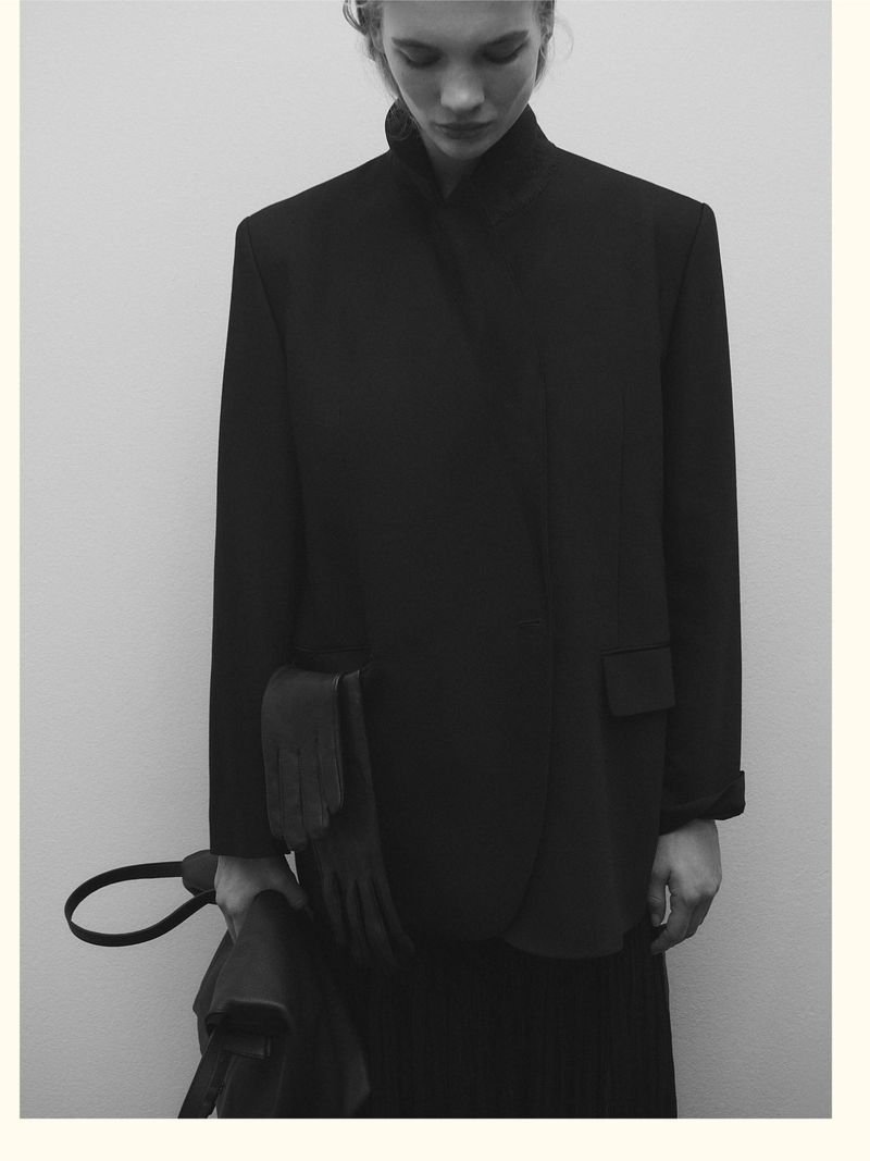 Model wearing a black structured blazer and holding a handbag against a neutral background