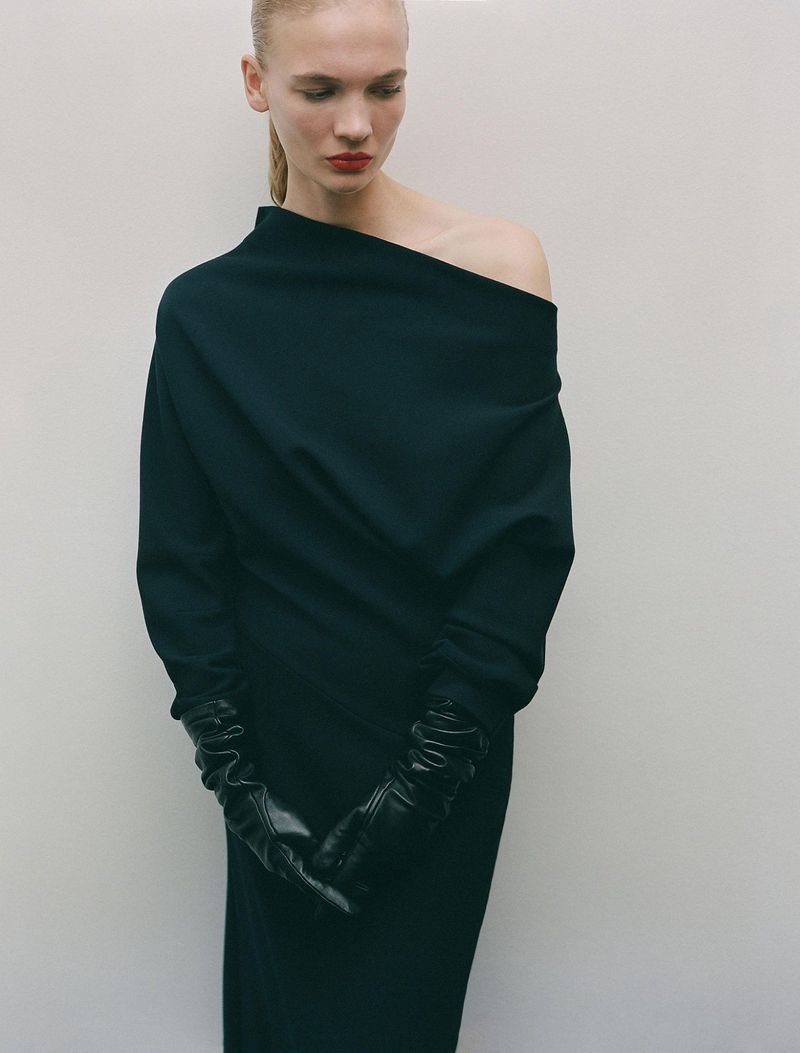 Model in a dark asymmetric dress wearing long black leather gloves against a plain light background