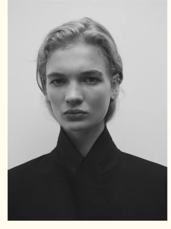 Close-up portrait of a woman with blonde hair wearing a black high-collared jacket against a plain grey studio backdrop.