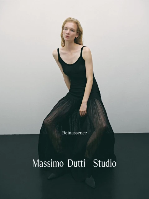 Fashion editorial shot of a blonde model in a black dress with pleated skirt, posing in a studio with a neutral background.