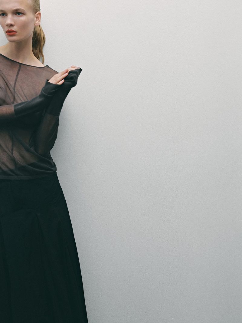 Model in sheer brown top and black skirt posing against a plain grey studio backdrop for Massimo Dutti