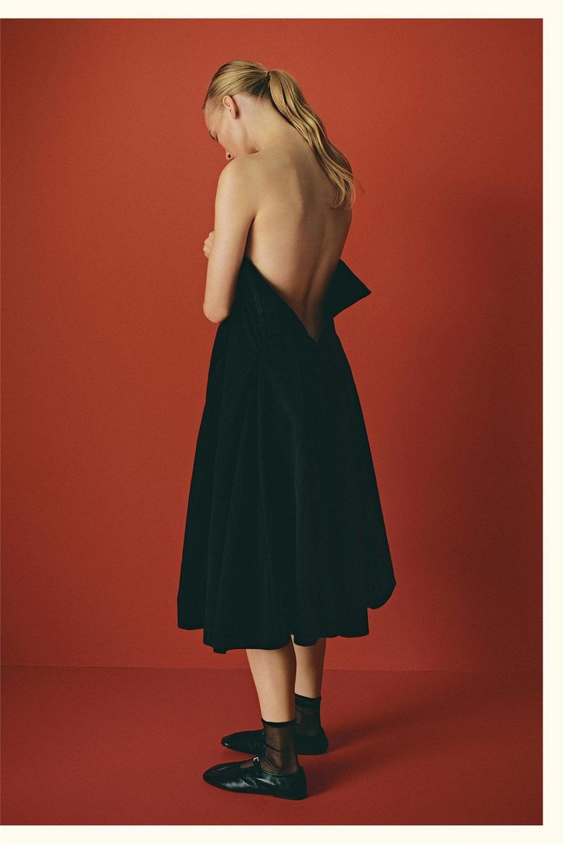 Blonde woman in black dress poses against red studio backdrop, showcasing fashion, style, and understated elegance in a minimalist setting.