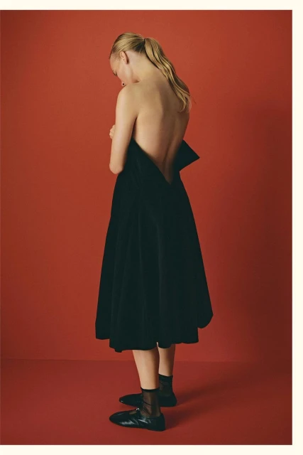 Blonde woman in black dress poses against red studio backdrop, showcasing fashion, style, and understated elegance in a minimalist setting.