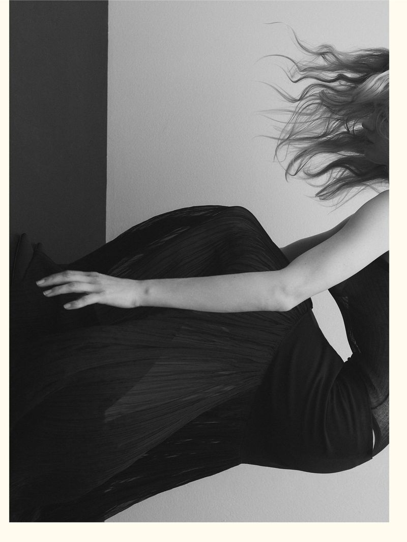 Monochrome fashion portrait of a model with flowing hair wearing a dark pleated skirt against a stark background