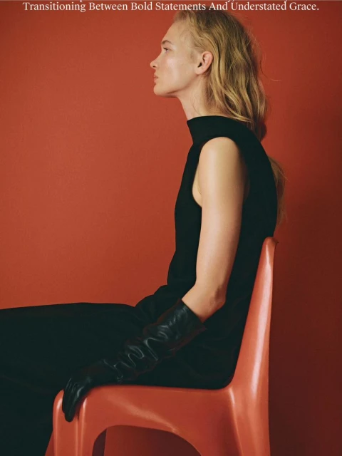 Elegant woman in black top and gloves seated against a terracotta backdrop, highlighted by soft, flattering light. Fashion editorial.