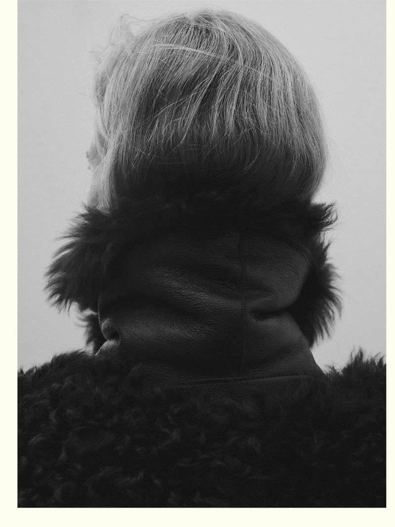 Monochrome portrait from behind features an adult with blond hair wearing a textured dark coat, set against a neutral grey background.