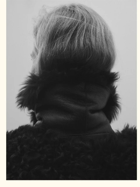 Monochrome portrait from behind features an adult with blond hair wearing a textured dark coat, set against a neutral grey background.