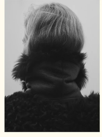 Monochrome portrait from behind features an adult with blond hair wearing a textured dark coat, set against a neutral grey background.