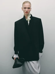 Model in oversized black blazer and white skirt holding a dark woven clutch against a seamless neutral background.