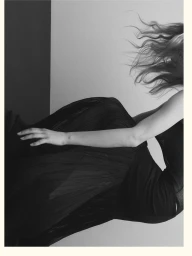 Monochrome fashion portrait of a model with flowing hair wearing a dark pleated skirt against a stark background