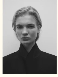 Close-up portrait of a woman with blonde hair wearing a black high-collared jacket against a plain grey studio backdrop.