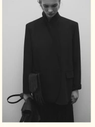Model wearing a black structured blazer and holding a handbag against a neutral background