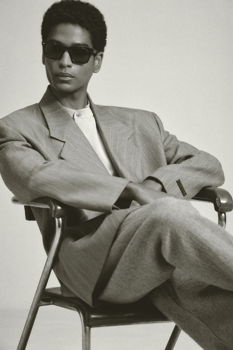 Stylish adult male model in sunglasses poses while seated in gray suit. Studio shot with neutral backdrop, medium shot, portrait orientation.