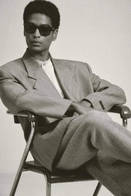 Stylish adult male model in sunglasses poses while seated in gray suit. Studio shot with neutral backdrop, medium shot, portrait orientation.