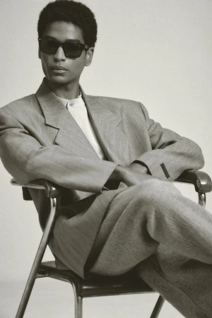 Stylish adult male model in sunglasses poses while seated in gray suit. Studio shot with neutral backdrop, medium shot, portrait orientation.