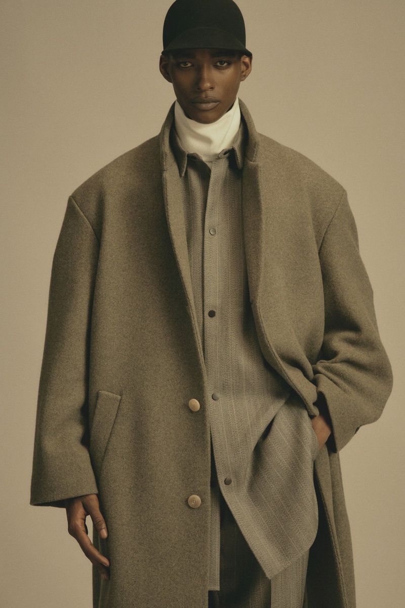 Fashion portrait of a young adult Black man in a wool coat, baseball cap, and suit in a neutral studio setting with soft lighting.